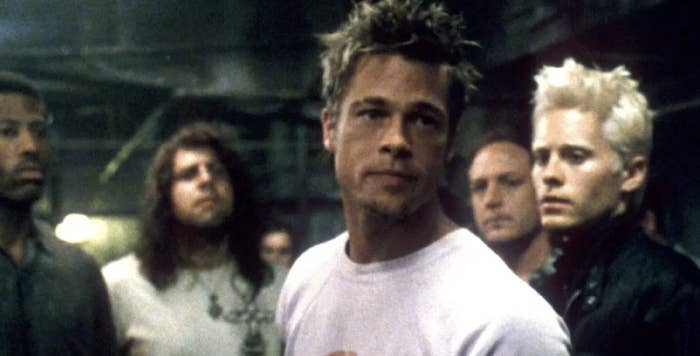 A group of men stand together in a dimly lit room, focusing on a central man with spiked hair and a white t-shirt, suggesting an intense scene