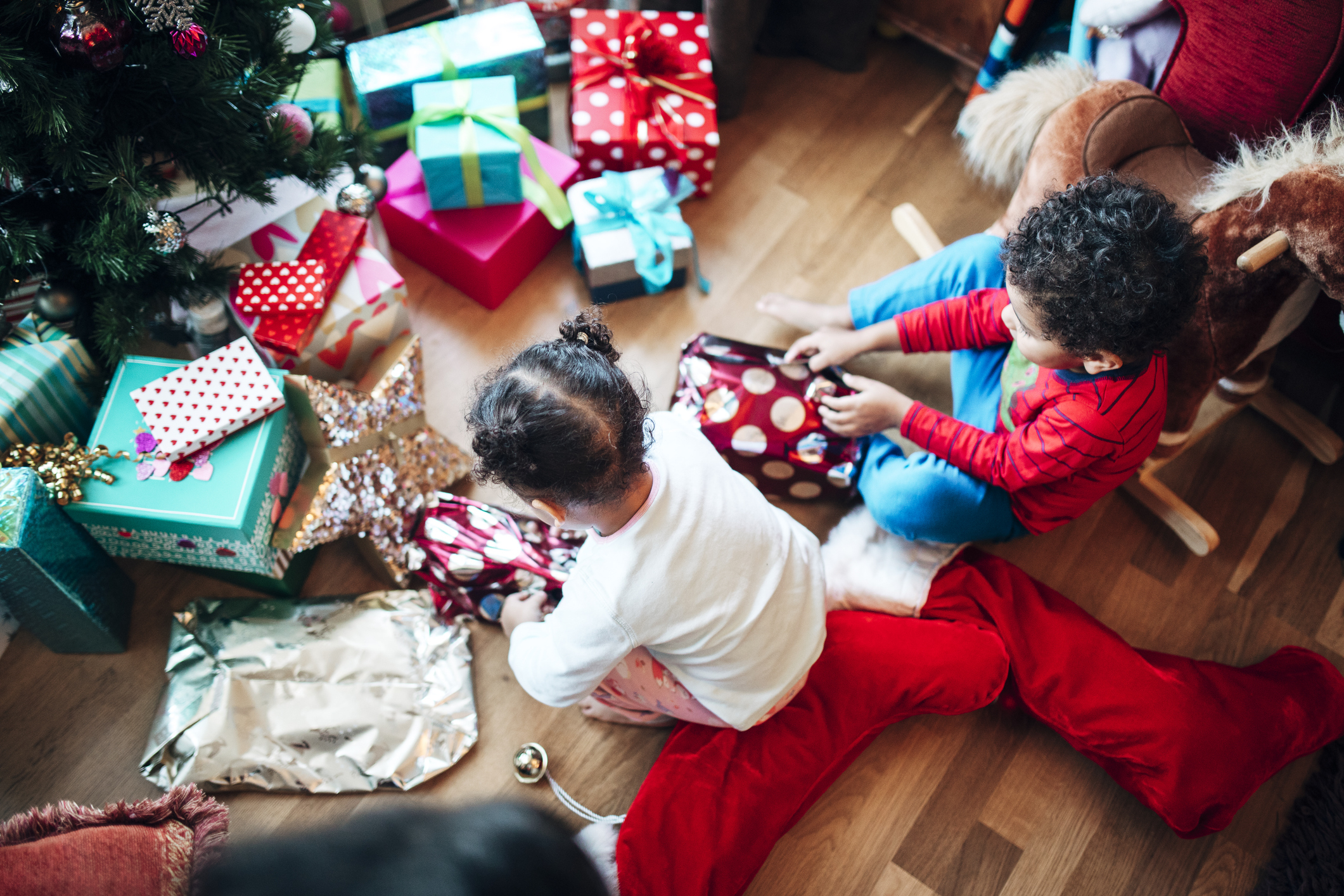 Children happily unwrapping gifts by a Christmas tree beside a large stocking and festive presents