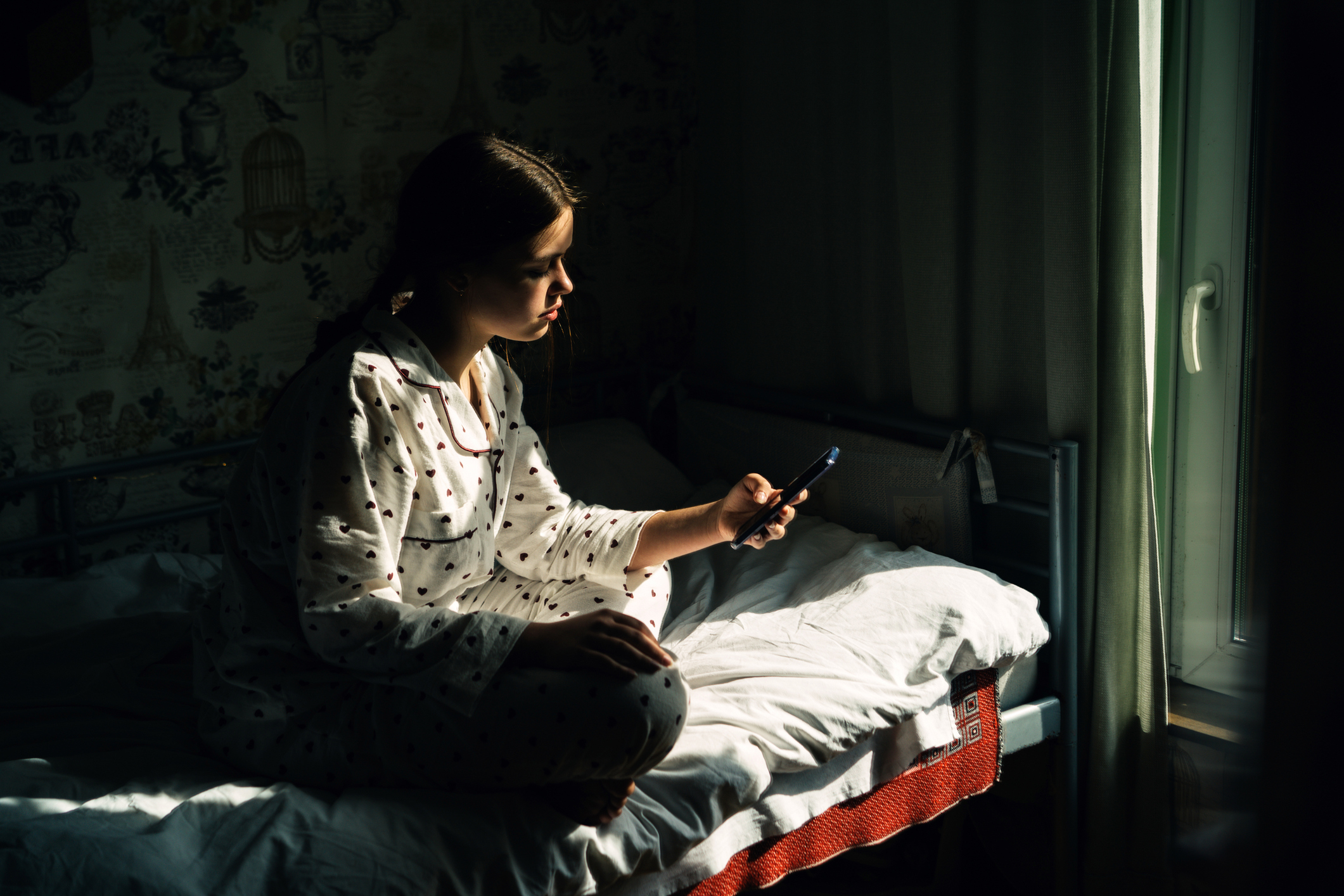 Person in patterned pajamas sits on a bed, using a smartphone near a window with soft light