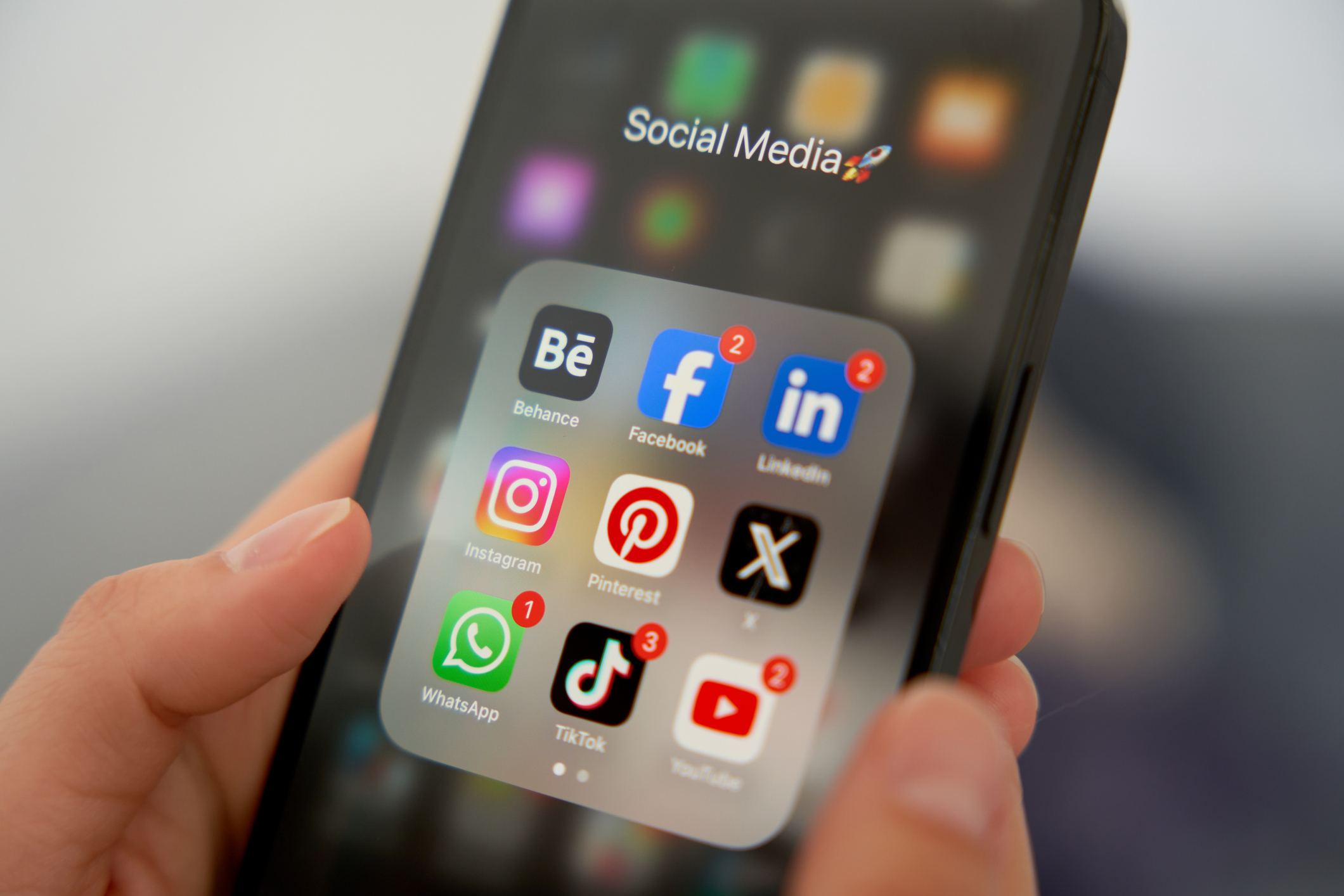 Person holding smartphone showing social media app icons including Facebook, Instagram, TikTok, and others, with notification badges on some