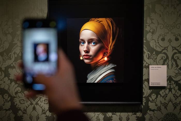 Painting of a woman with a yellow headscarf and pearl earring; a blurred hand holding a phone in the foreground