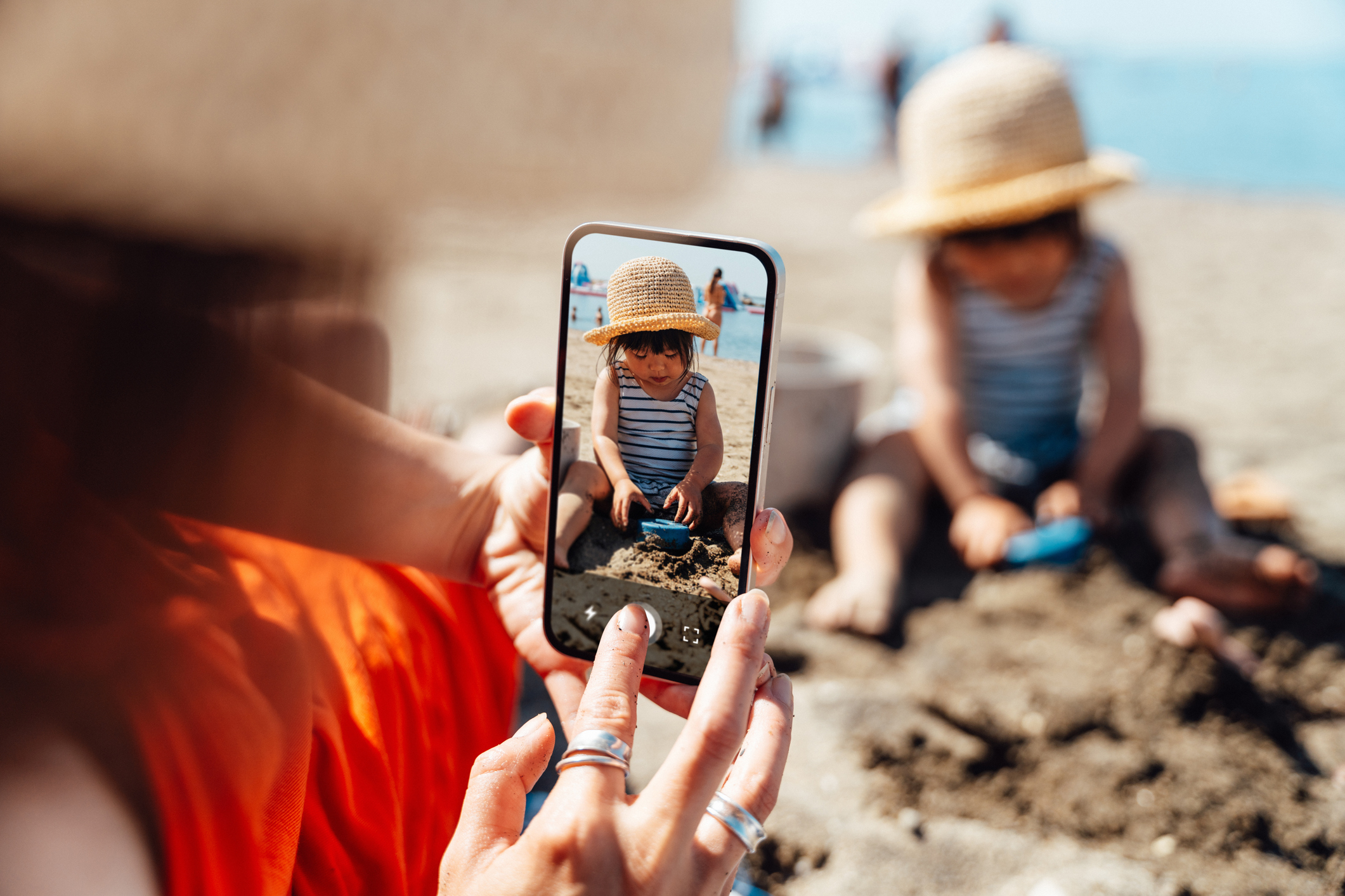 Person taking a photo of a child playing on a sandy beach, captured on a smartphone screen. Both wear hats; the child is focused on the sand