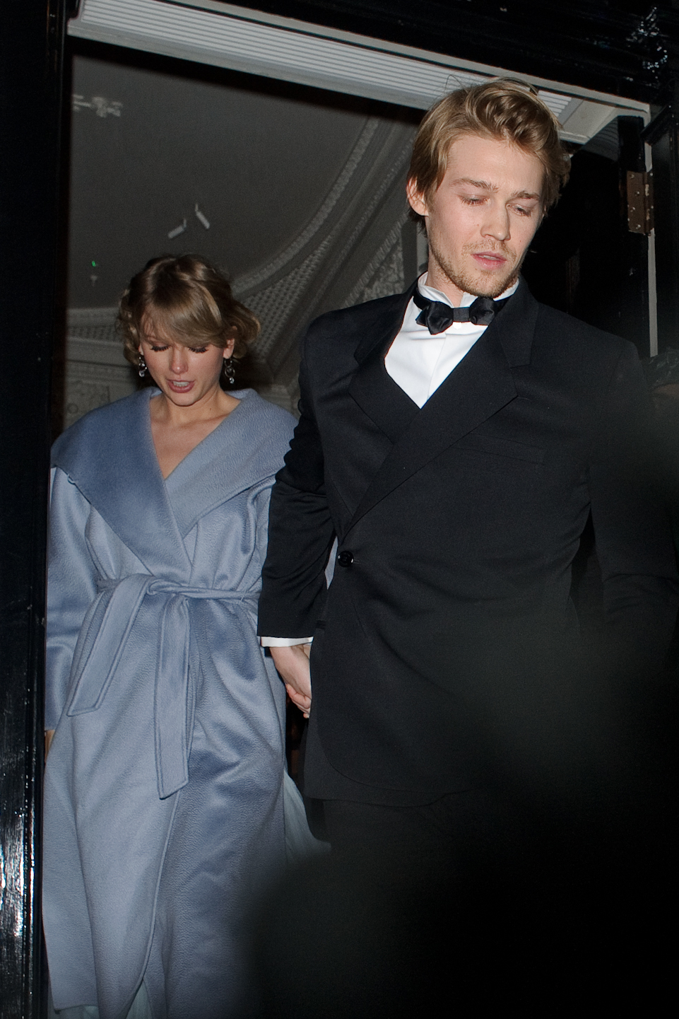 Two celebrities leaving an event; one wears a belted coat, the other a tuxedo with a bow tie