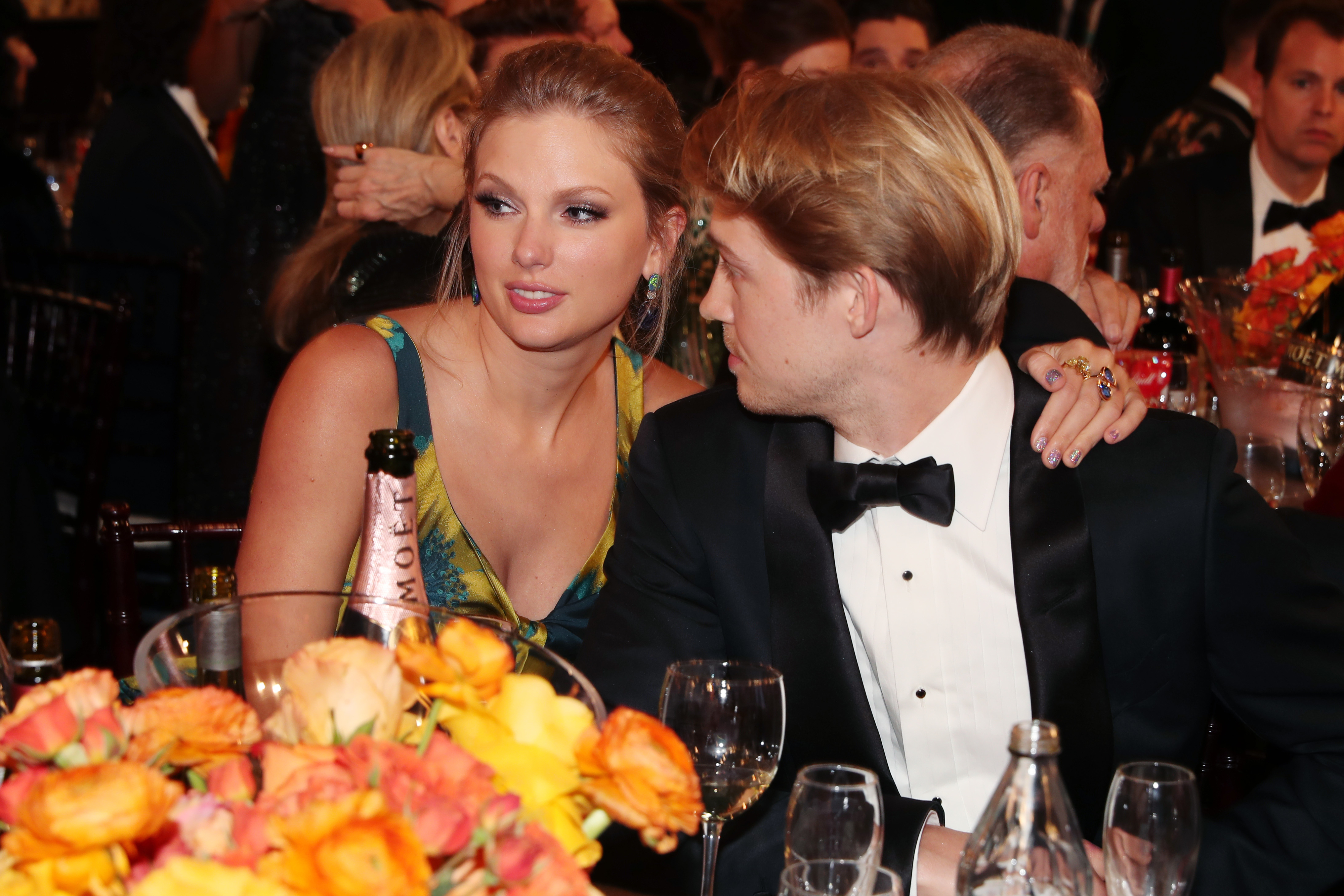 Two people sit closely at a formal event, surrounded by orange and yellow flowers. The woman wears a floral dress, the man in a tuxedo looks away