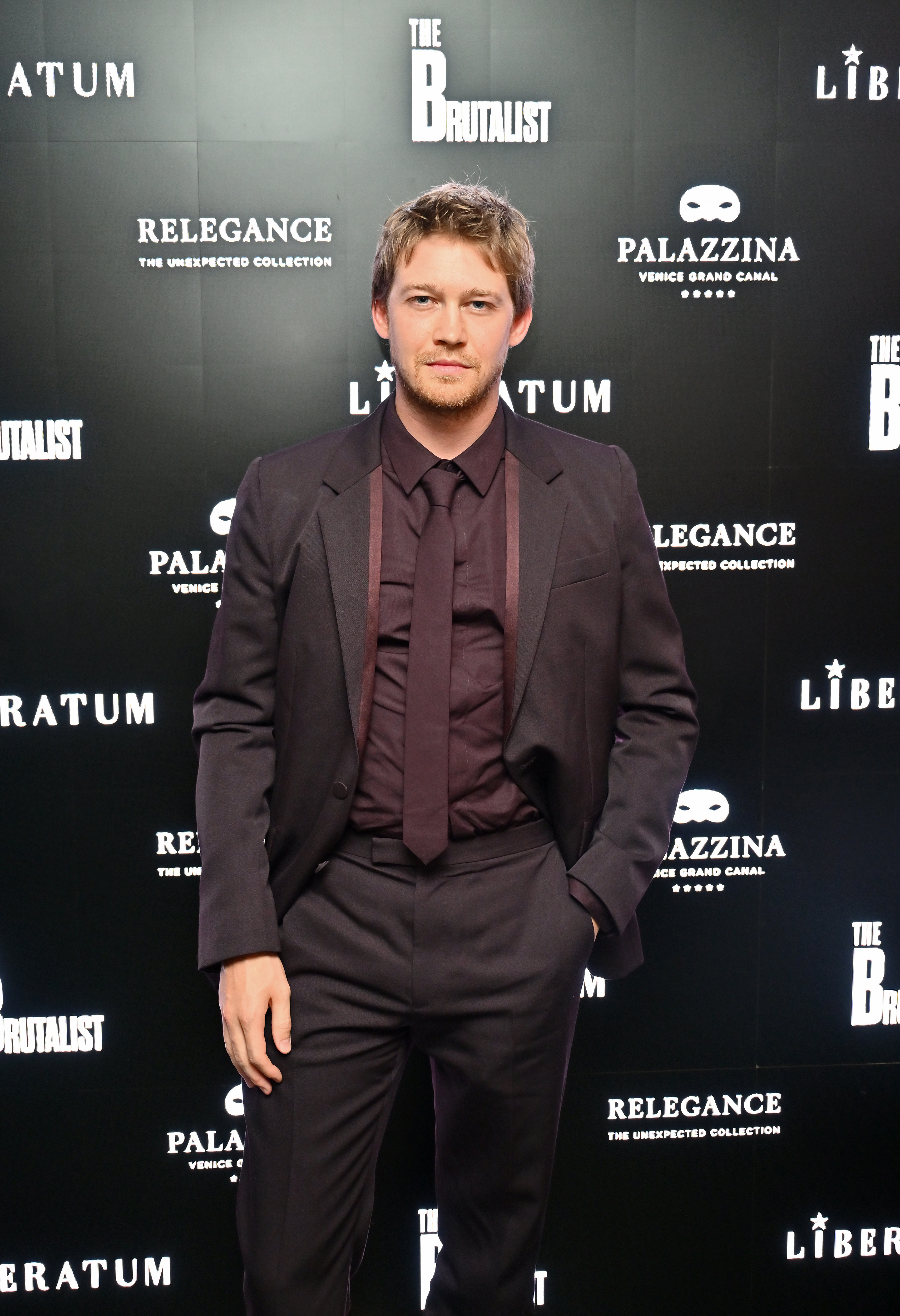 Joe Alwyn in a tailored suit with a tie poses on a red carpet with &quot;The Brutalist&quot; signs in the background