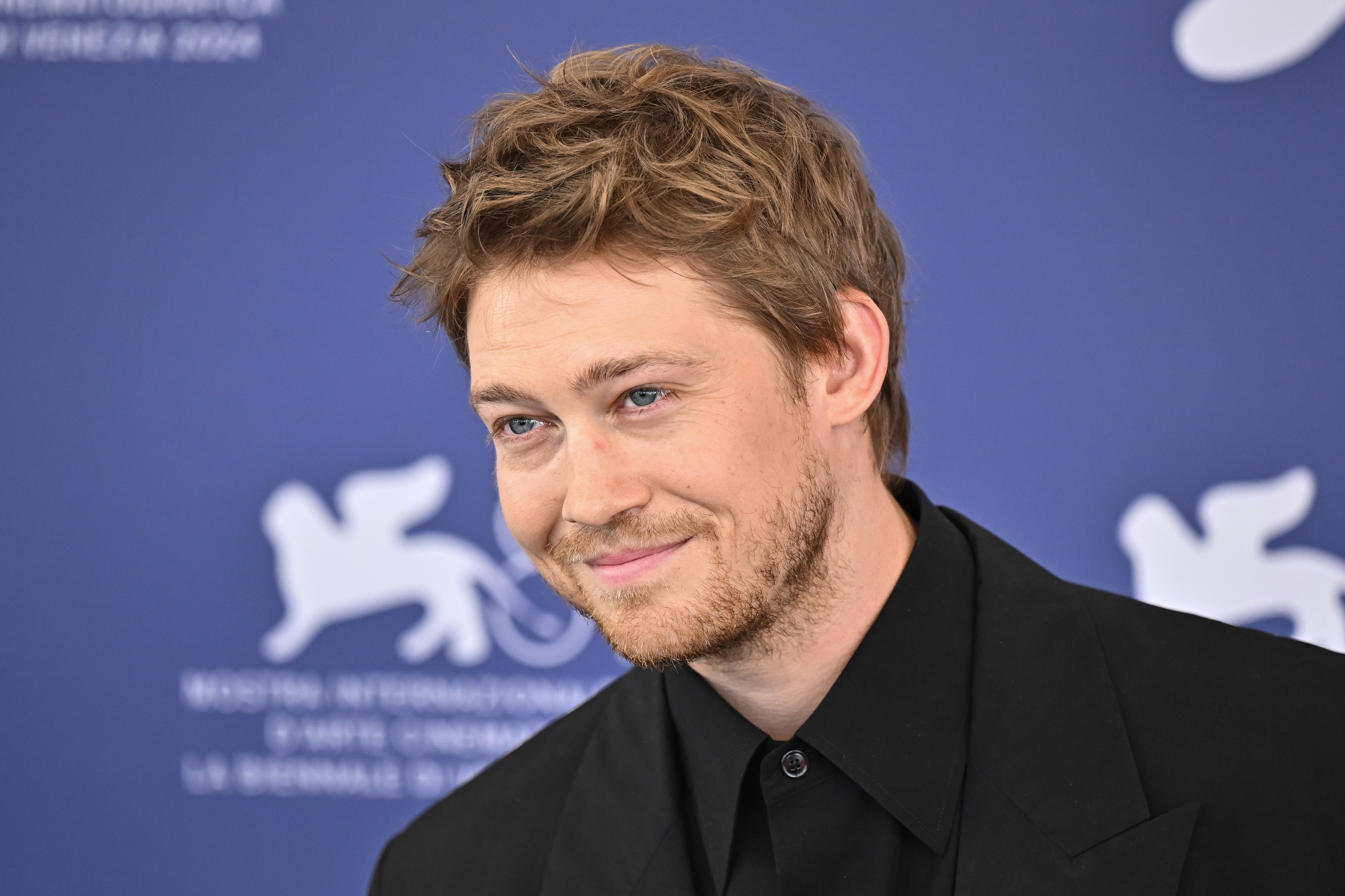 Joe Alwyn in formal attire at a film festival press event, smiling against a backdrop with festival logos