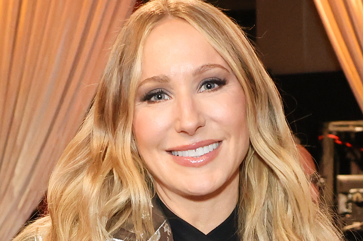 Nikki Glaser wearing a metallic suit with a cut-out top, posing indoors, smiling with a background of draped curtains and tables