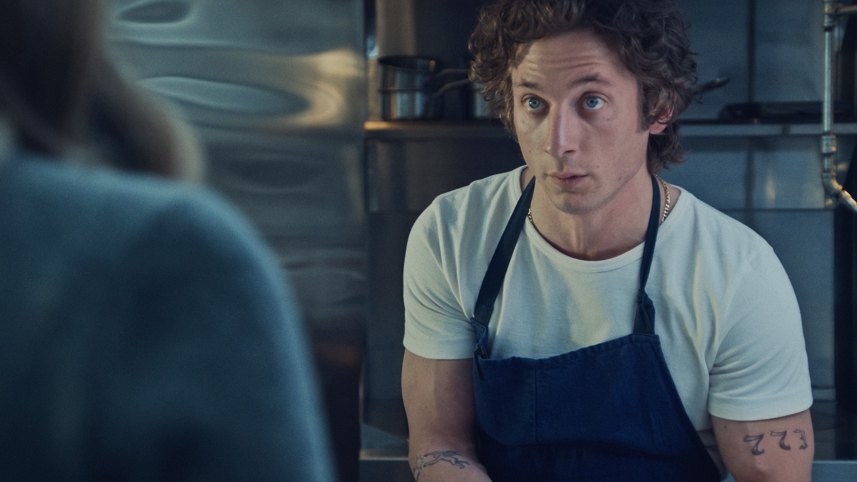 A person with curly hair in a kitchen, wearing a white t-shirt and dark apron, looks intently at someone off-camera