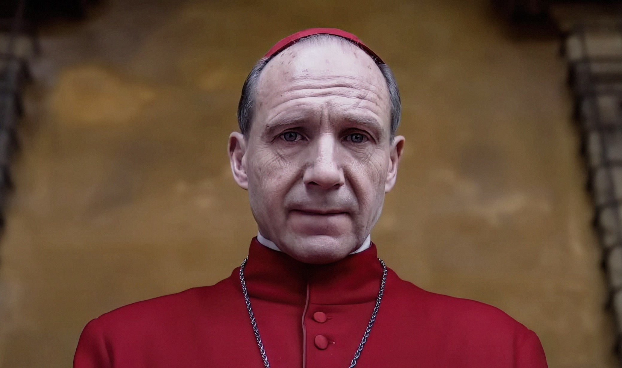 A serious-looking man in a red cardinal robe stands in front of a textured wall, wearing a red zucchetto and a chain necklace