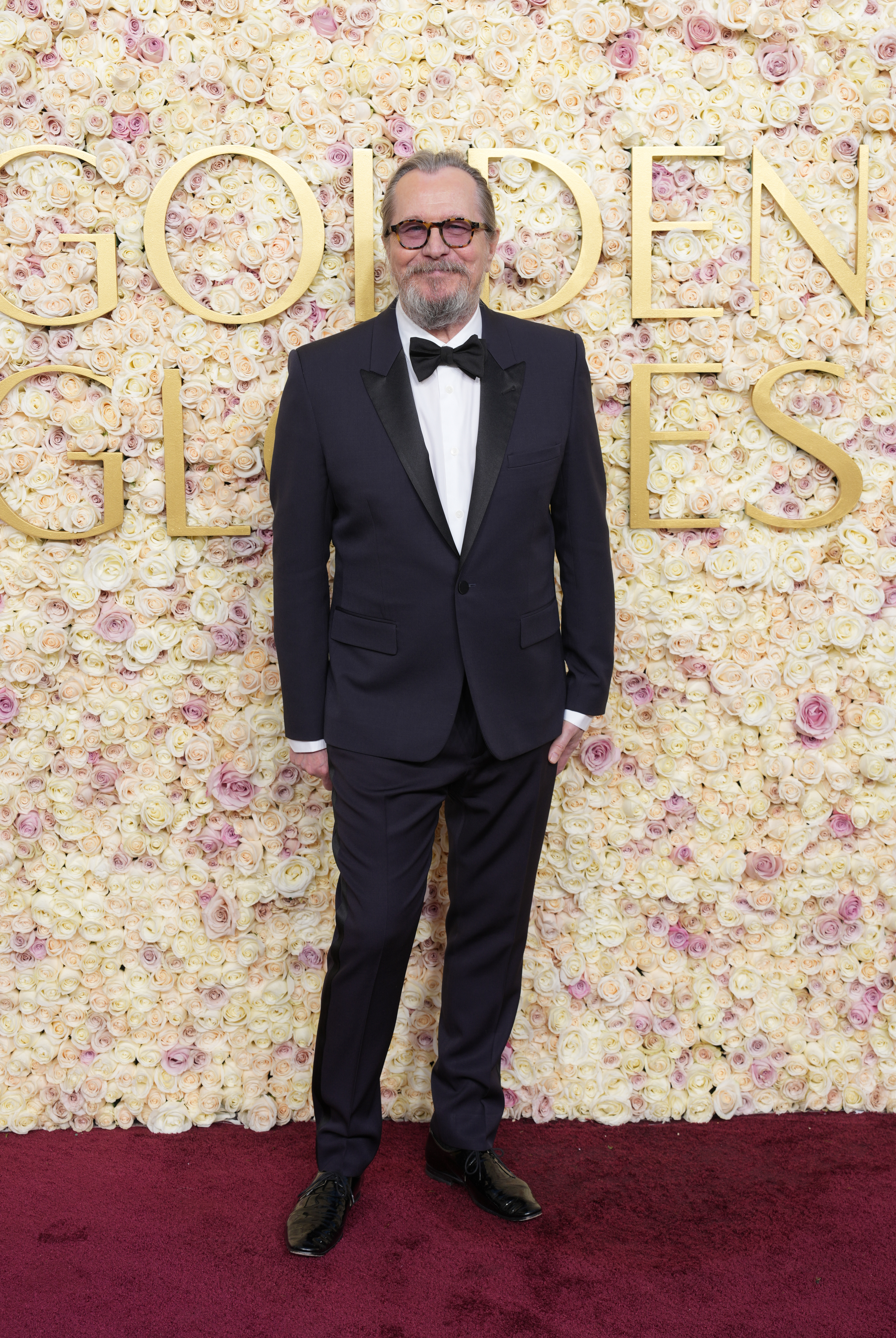 Gary Oldman in a black tuxedo with a bow tie stands on a red carpet in front of a floral wall with &quot;Golden Globes&quot; partially visible