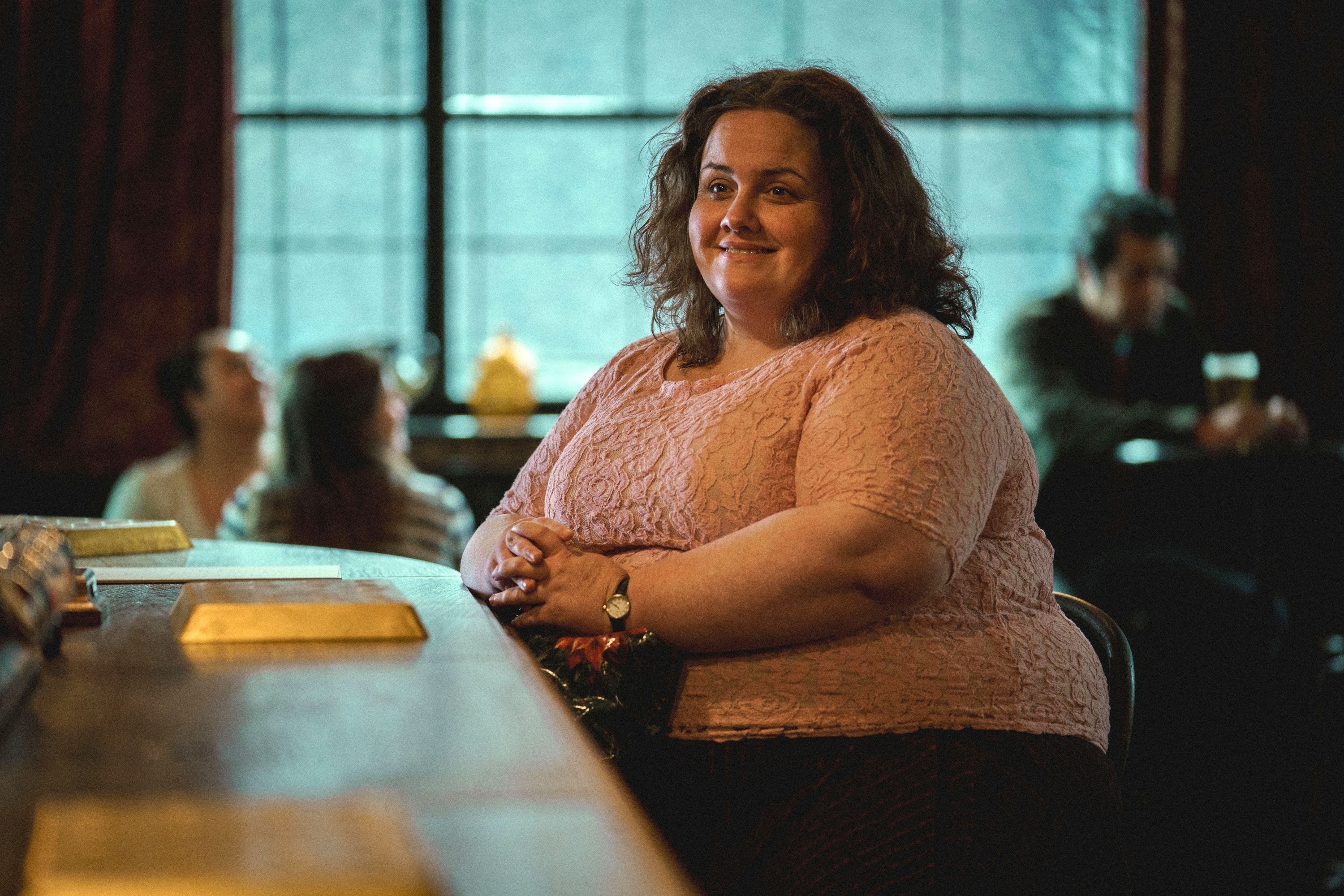 A woman sits at a bar, smiling, with three people in the blurred background, creating a warm and relaxed atmosphere