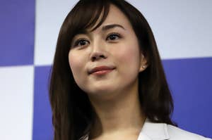 女性が微笑んでカメラを見つめている。彼女はフォーマルなジャケットを着ており、背景には広告用のボードが見える。