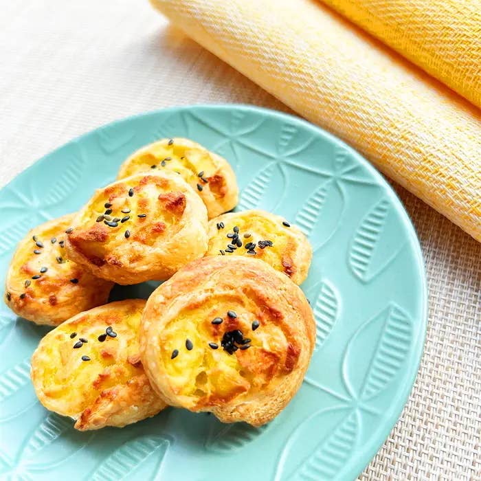 皿にチーズと黒ゴマの乗った焼きたてのペストリーが載っている。背景に黄色の布。
