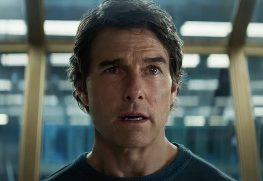 A person with short hair and a focused expression looks slightly upward indoors, standing in front of a blurred background