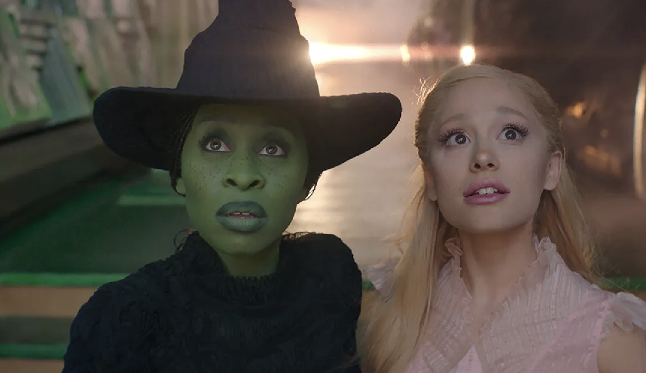 Two characters from a musical, one with green skin in a witch's hat and dark outfit, the other with light skin and long hair in a light dress, looking up