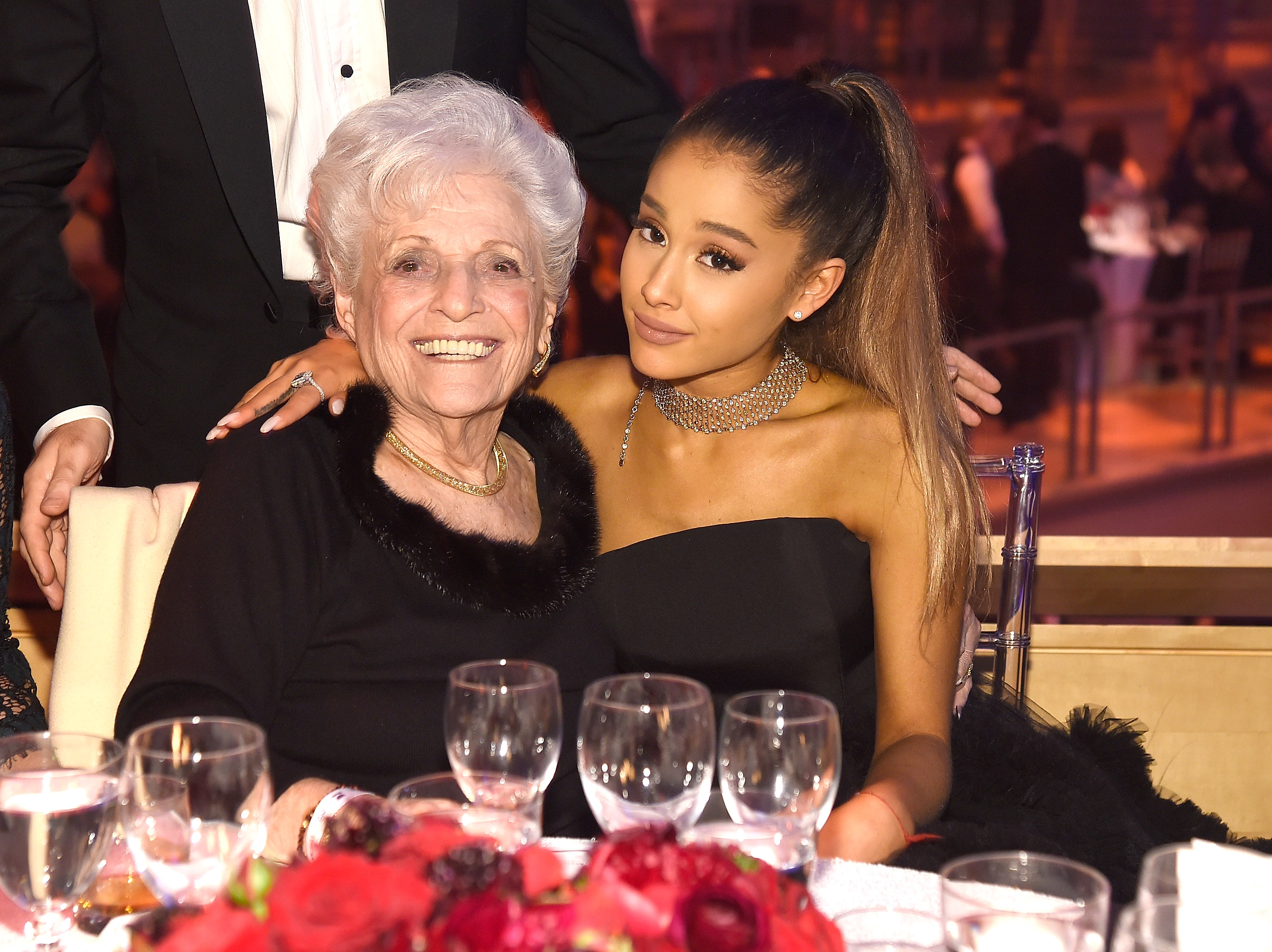 Ariana Grande and her Nonna seated at a formal event, Ariana wearing a strapless dress and choker. Both are smiling at the camera