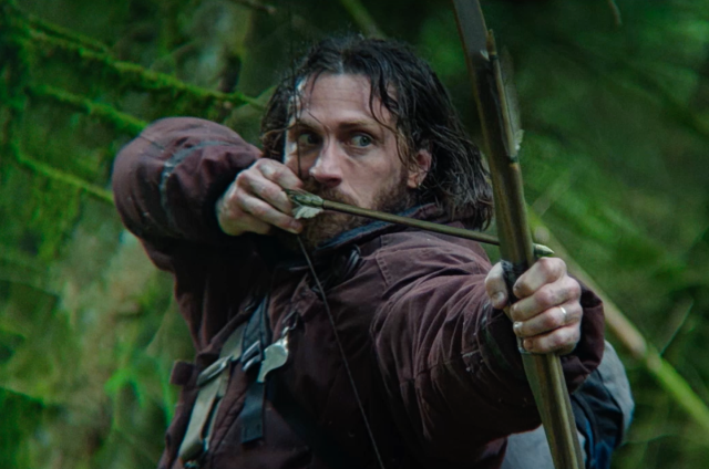 Person outdoors in rugged clothing, aiming a longbow with a focused expression in a forest setting