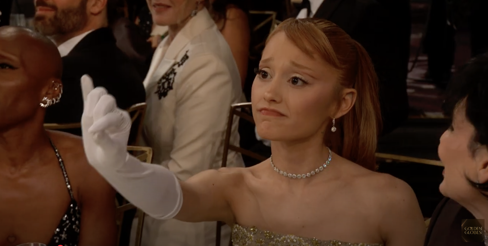 Person at an event wears elegant strapless gown and white gloves, gesturing with a finger raised. Other attendees are seated nearby