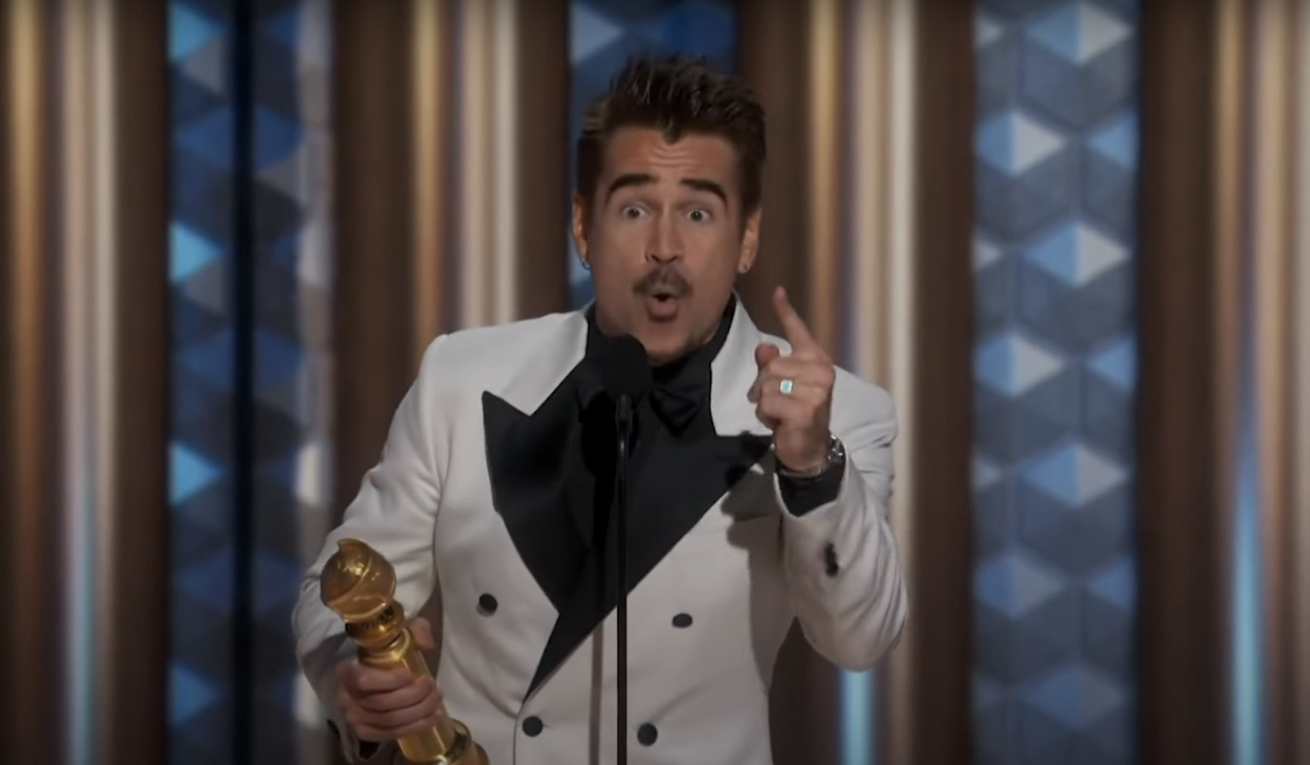 Colin Farrell in formal tuxedo holding an award on stage, speaking into a microphone with a surprised expression