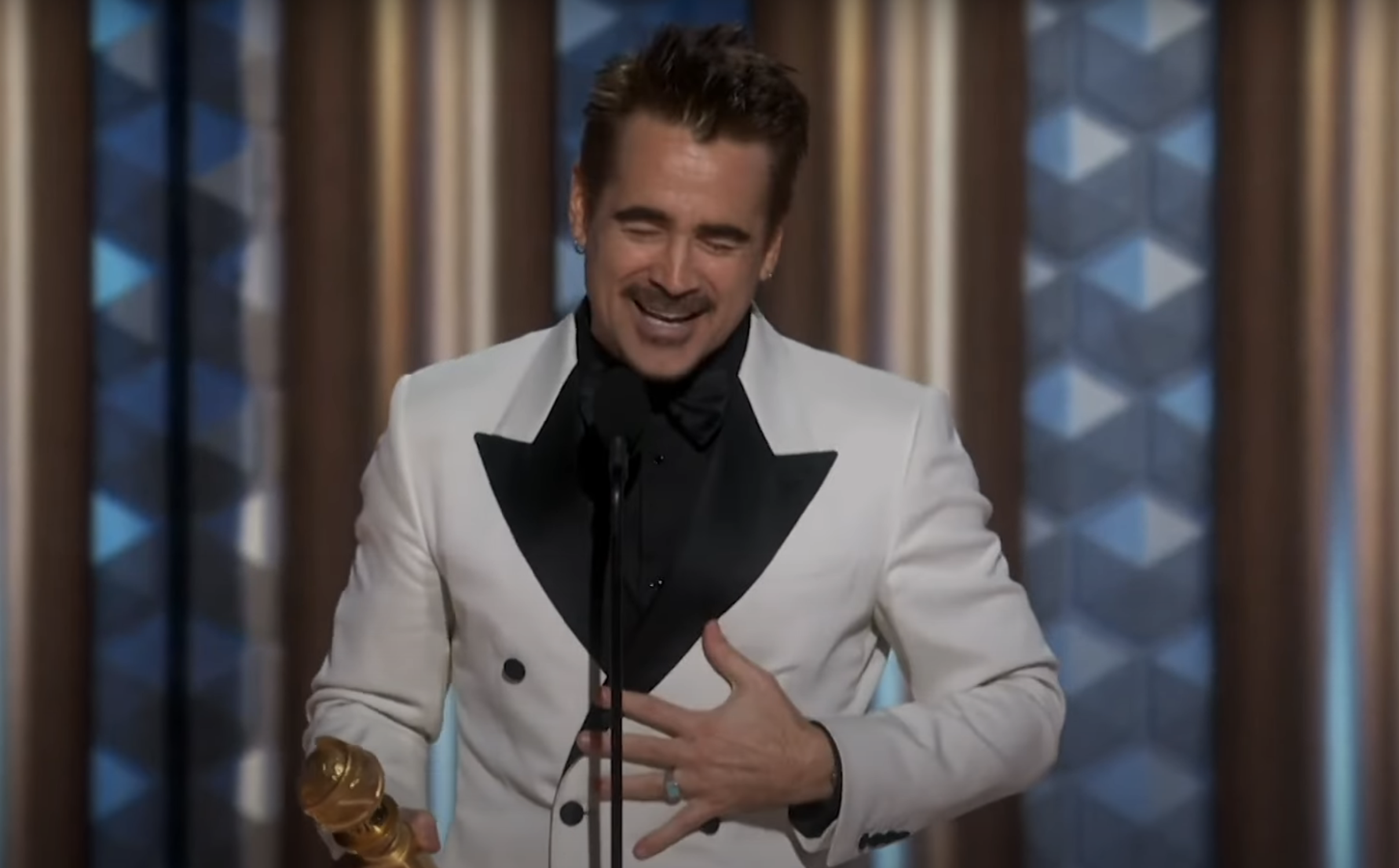 Colin Farrell in a tuxedo with a double-breasted white jacket holds a trophy, smiling and speaking at an event