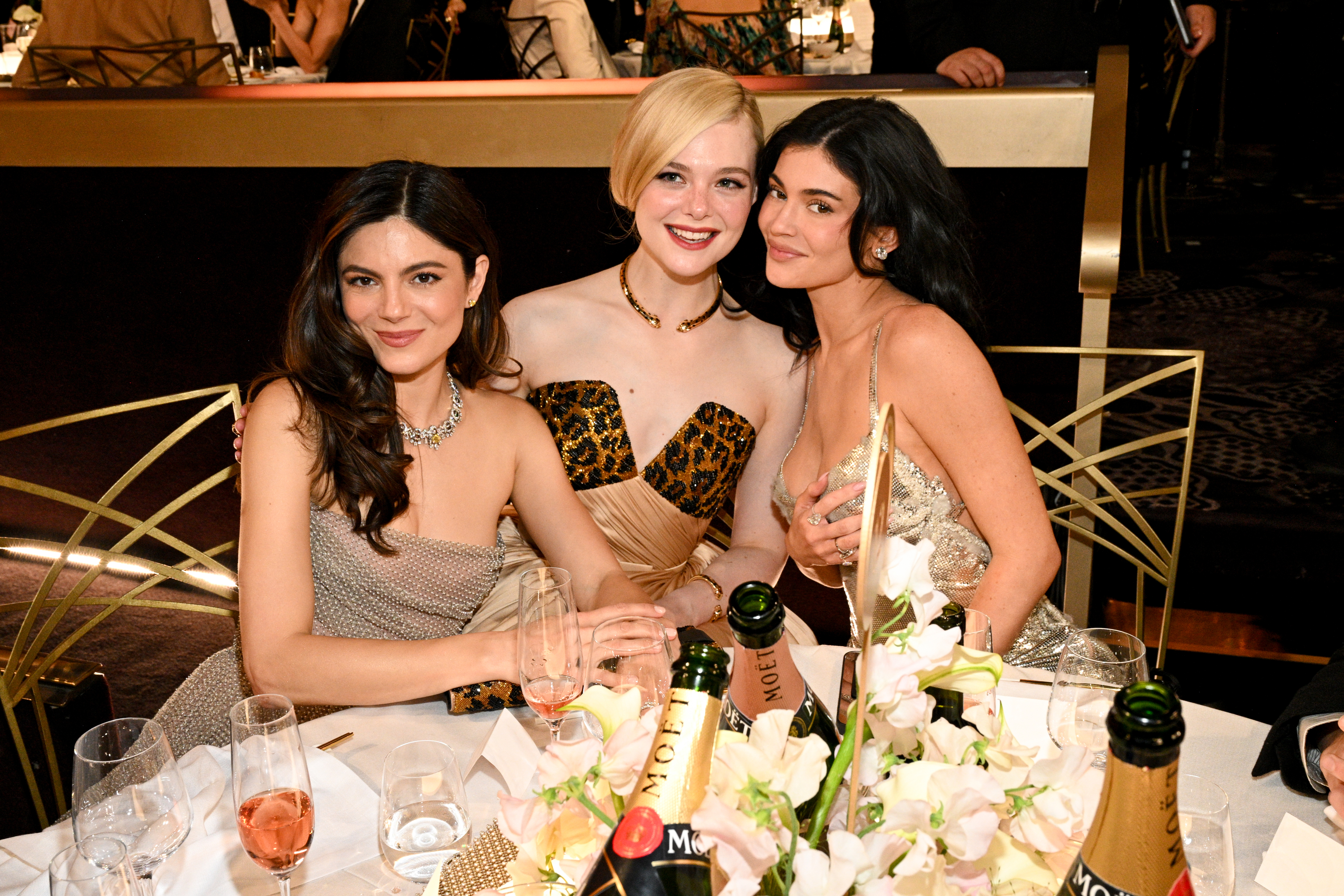 Three women seated at a formal event table, smiling and dressed in elegant evening gowns