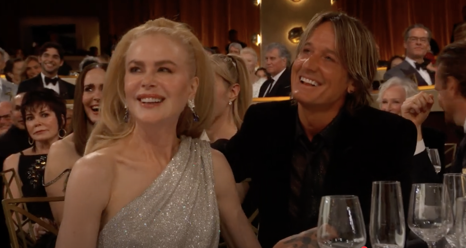 Two people in evening attire sit smiling at an event, surrounded by other attendees at tables