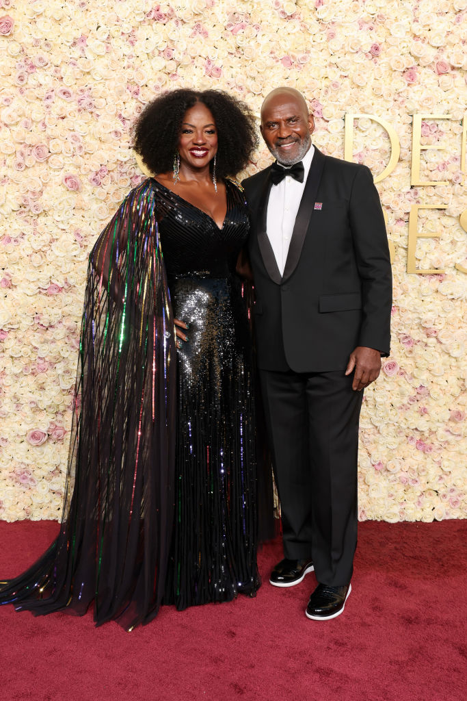 The couple pose on a red carpet. Viola wears a shimmering gown with a cape, and the man is in a classic tuxedo