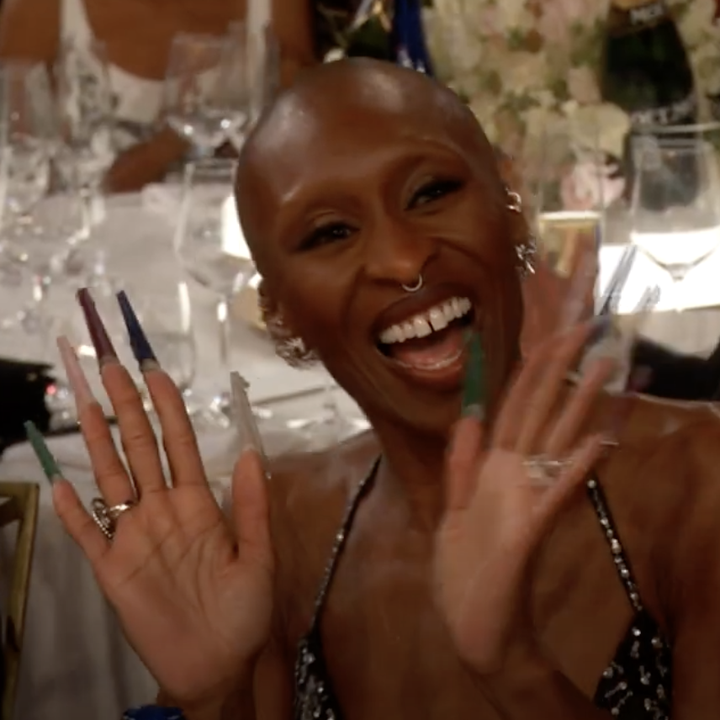Person smiling and waving at an event table, dressed in a patterned gown, surrounded by other attendees and formal dinner settings