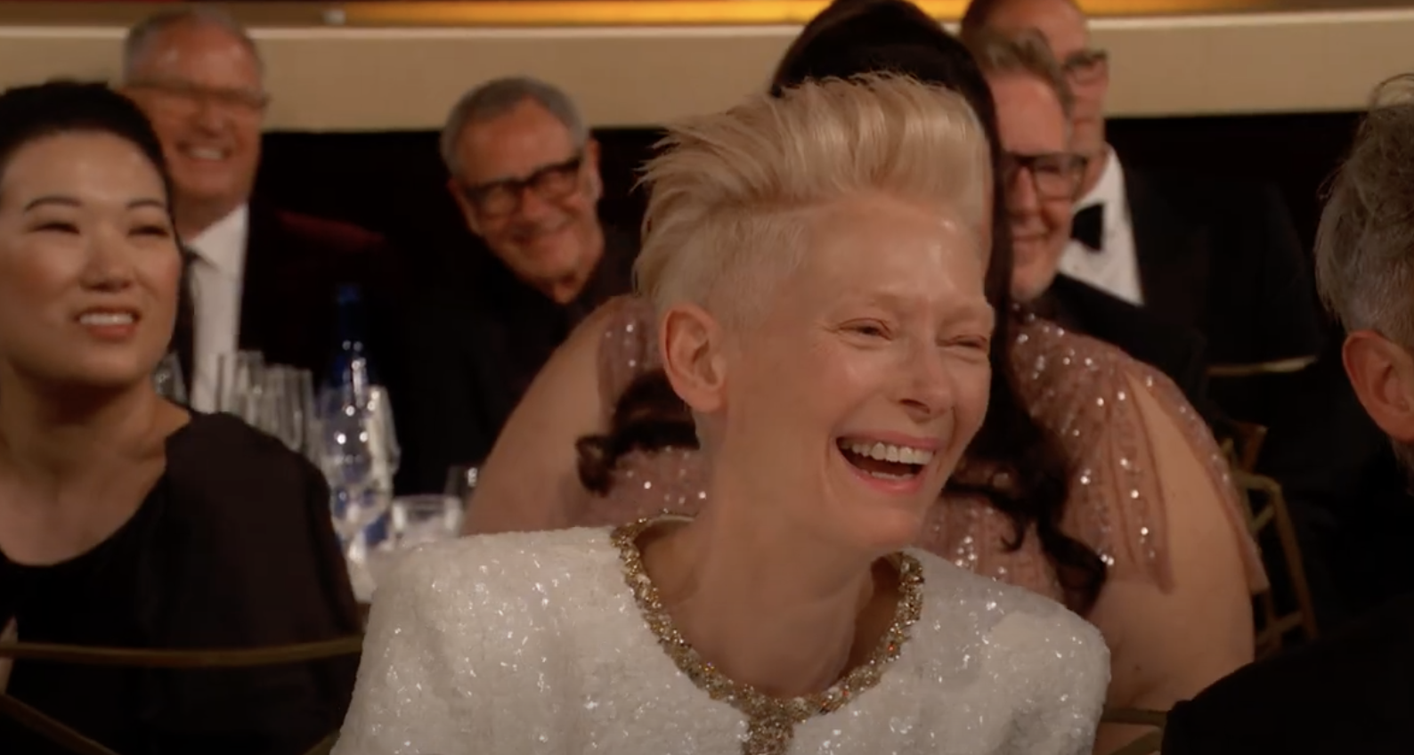 Person with short hair laughing, wearing a white textured dress with a jeweled neckline, seated at an event with others in formal attire