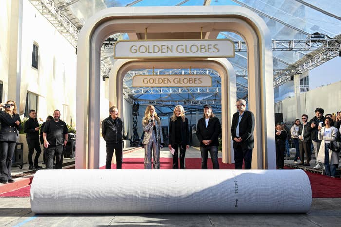 Group of people standing on a red carpet under a sign reading &quot;Golden Globes,&quot; with a large rolled-up carpet in the foreground