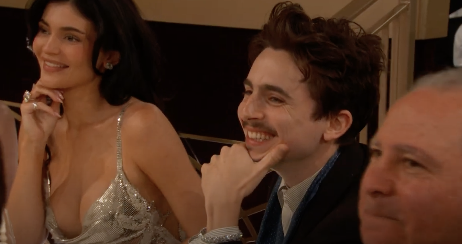 Two people smiling at an event, seated at a table. One is wearing a glittery dress, while the other wears a suit with a tie