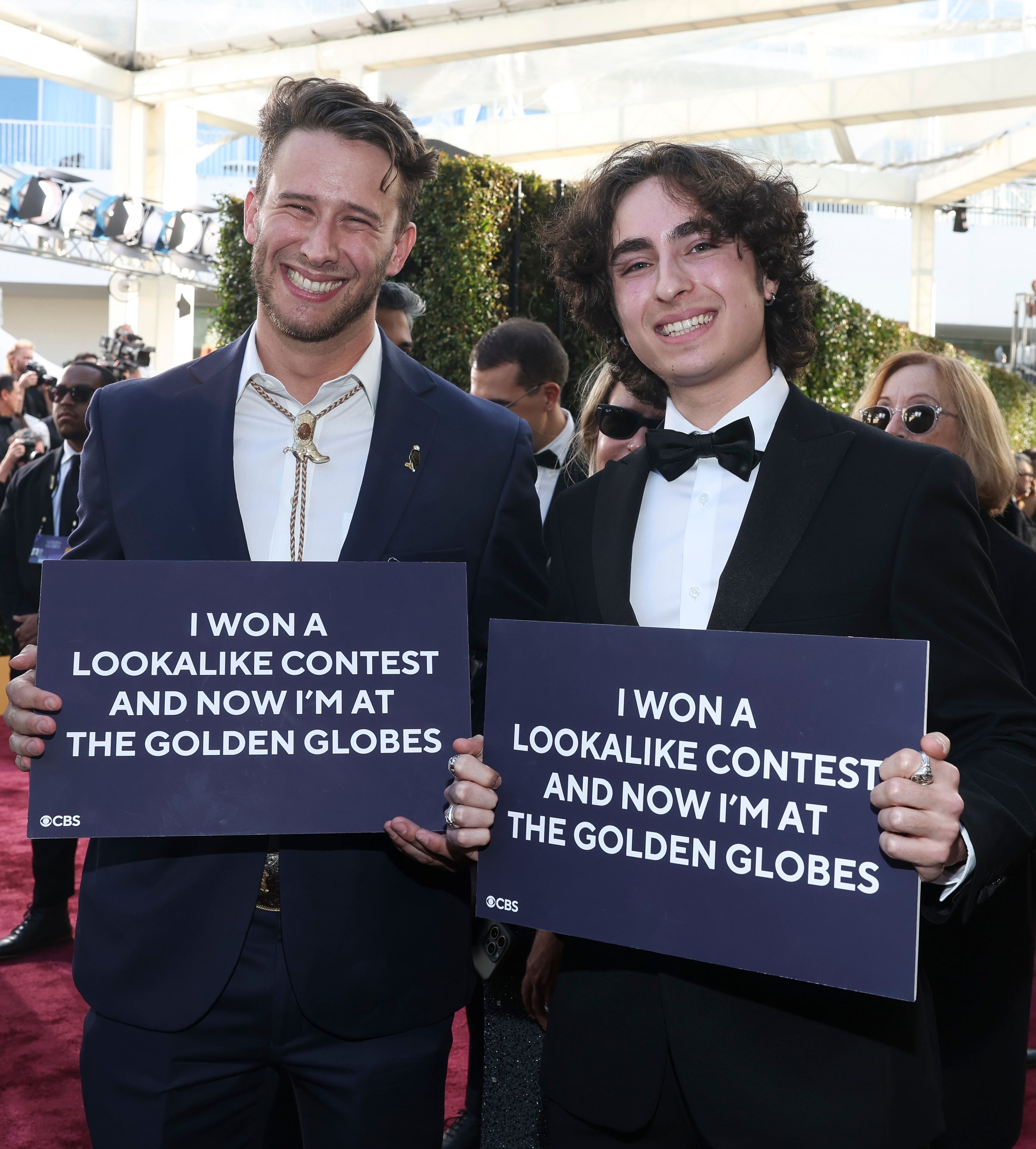 Two men holding signs saying they won a lookalike contest and are now at the Golden Globes, standing on the red carpet in formal attire