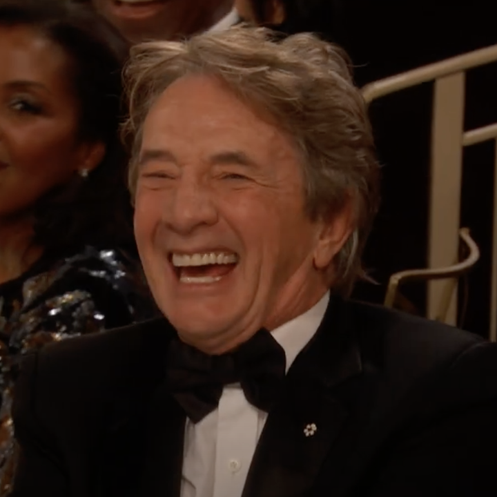 A person in a formal tuxedo laughs heartily while seated at an event, with others around enjoying the moment