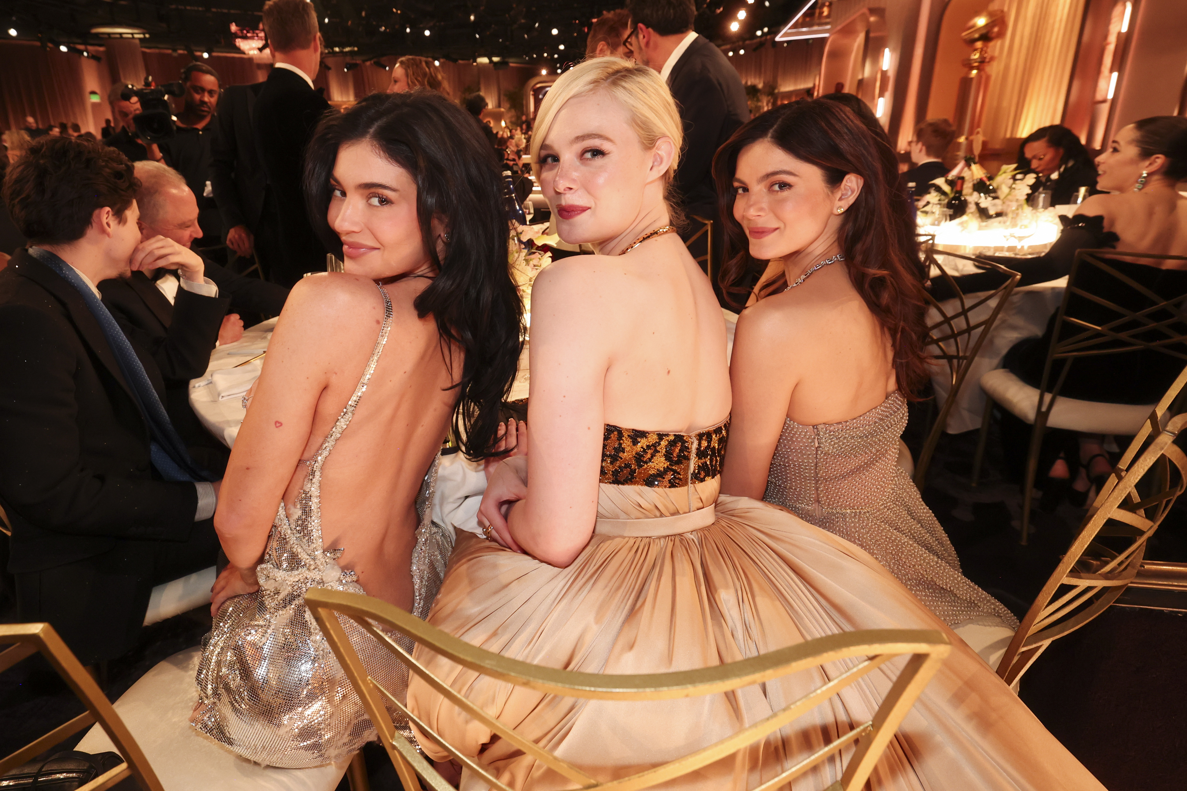 Three women seated at an elegant event, wearing formal gowns with intricate back designs, posing for a photo at a table