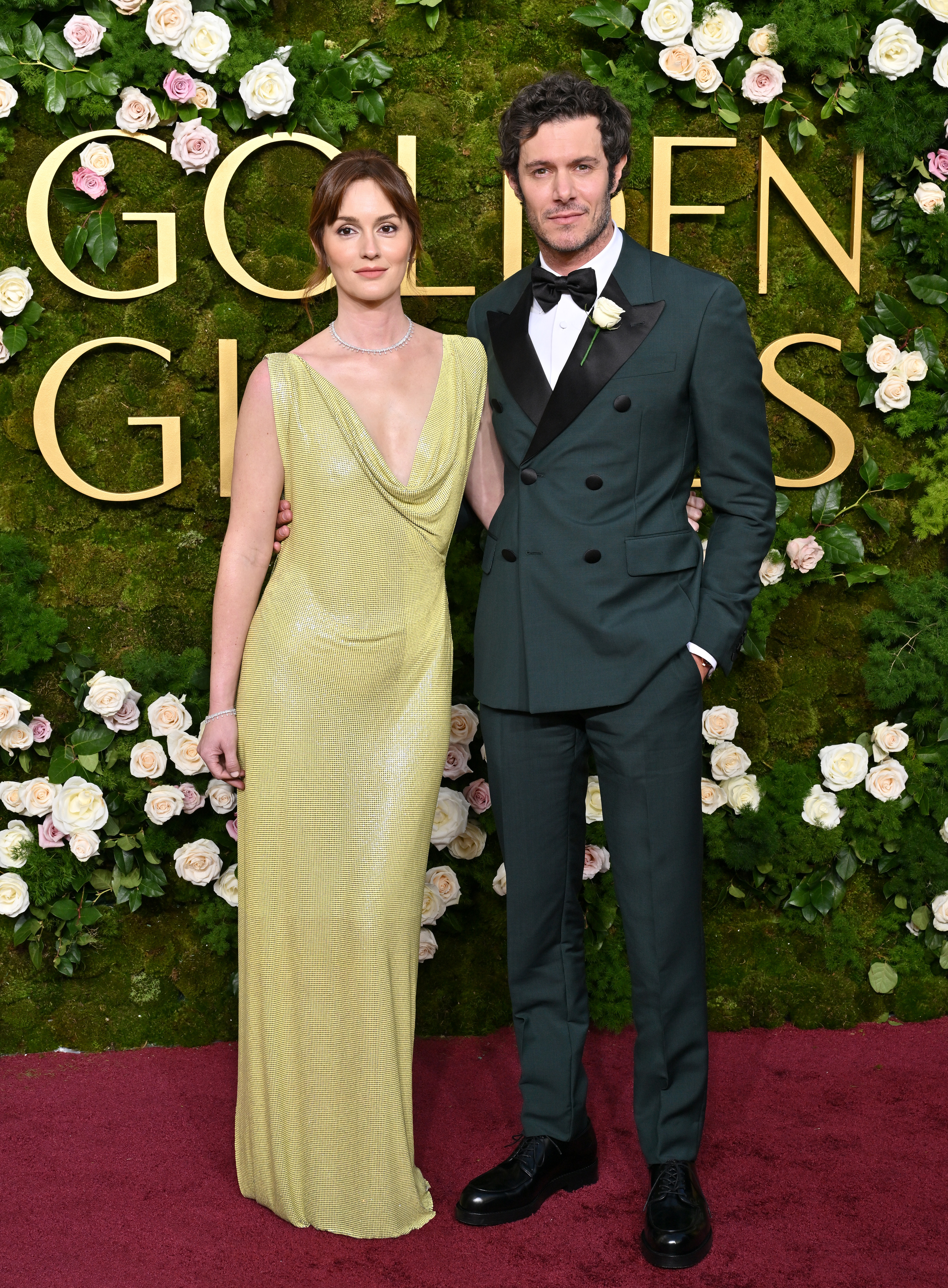 Two individuals on a red carpet; one in a shimmering gown, the other in a tailored suit, standing in front of a floral backdrop with the word &quot;GOLDEN.&quot;