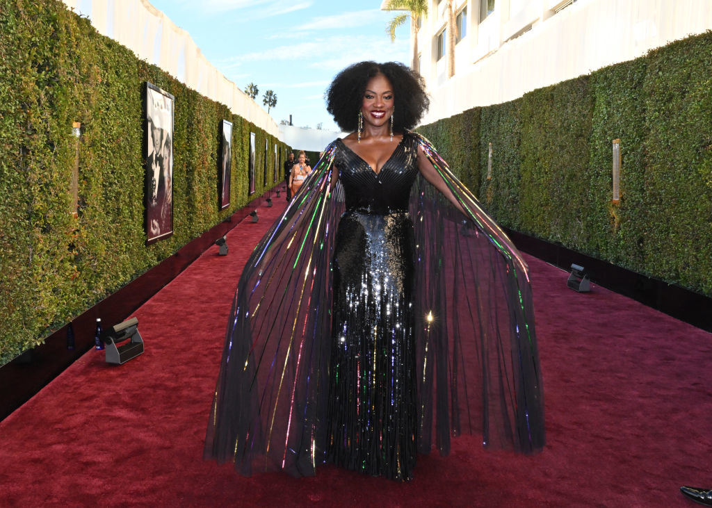 Woman on red carpet wearing a sparkling floor-length gown with sheer cape sleeves, smiling confidently