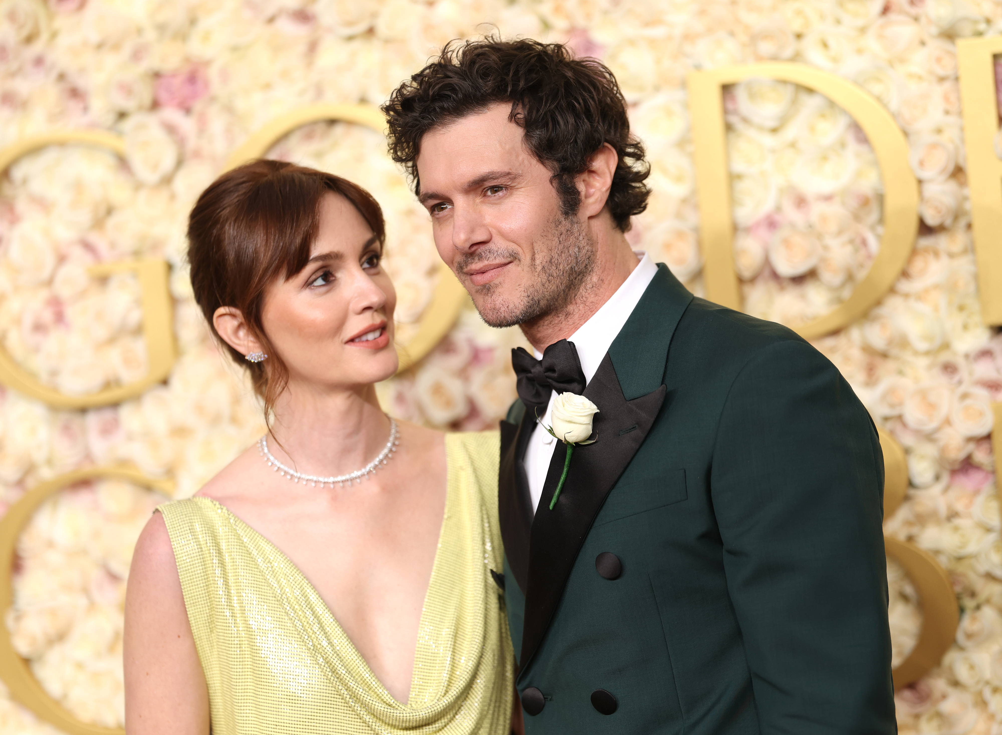 Adam in a tuxedo with a rose boutonniere and Leighton in an elegant sleeveless gown pose on a floral-themed red carpet