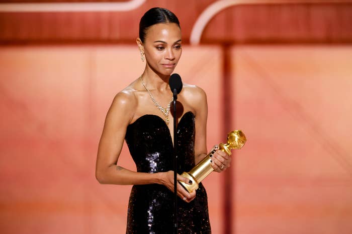 Zoe Saldaña in an elegant gown holds a Golden Globe award while speaking at a microphone on stage