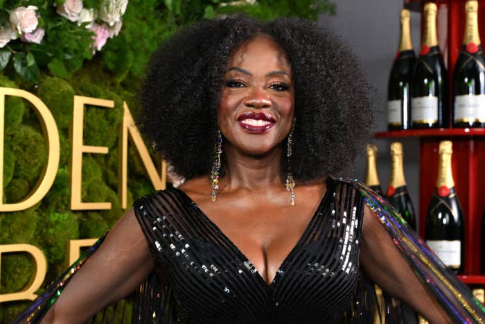 Viola on the red carpet wearing a gown with shimmering details, smiling. Background includes flowers and champagne bottles