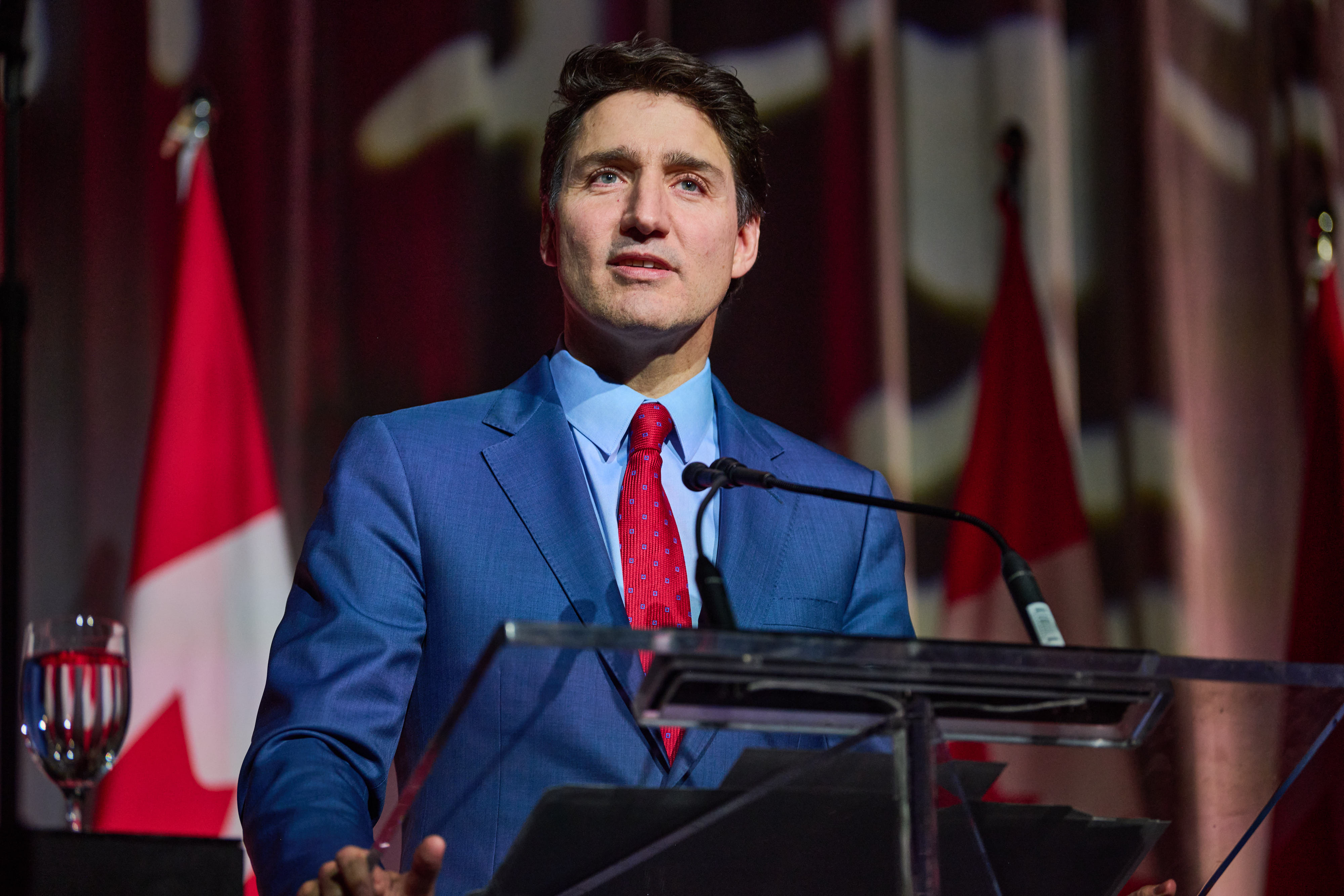 Person in a blue suit and red tie speaks at a podium with microphones. Canadian flags are visible in the background