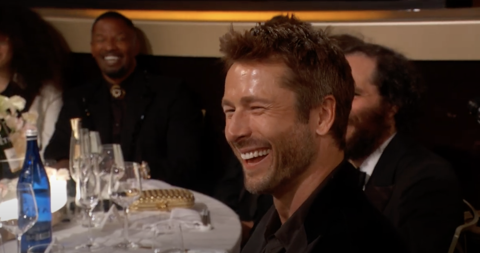 A man in formal attire laughing while seated at a table during a celebrity event. Other attendees are visible in the background