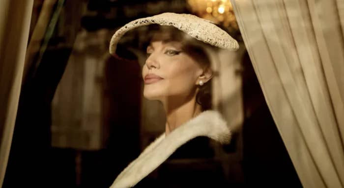 Woman in elegant hat with lace detail, looking over her shoulder, wearing a fur-collared coat, standing by curtains with a chandelier in the background