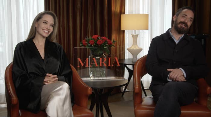 Two people seated, one in a black robe-style top, smiling, next to a man in a jacket, with a table holding roses and &quot;Maria&quot; between them