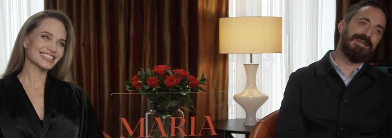 Two people sit smiling in a room with a sign reading "MARIA" on a table with red roses between them