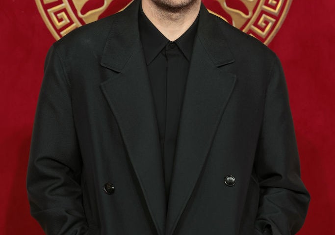 Sabato De Sarno in a dark, tailored overcoat on a red carpet with a decorative backdrop