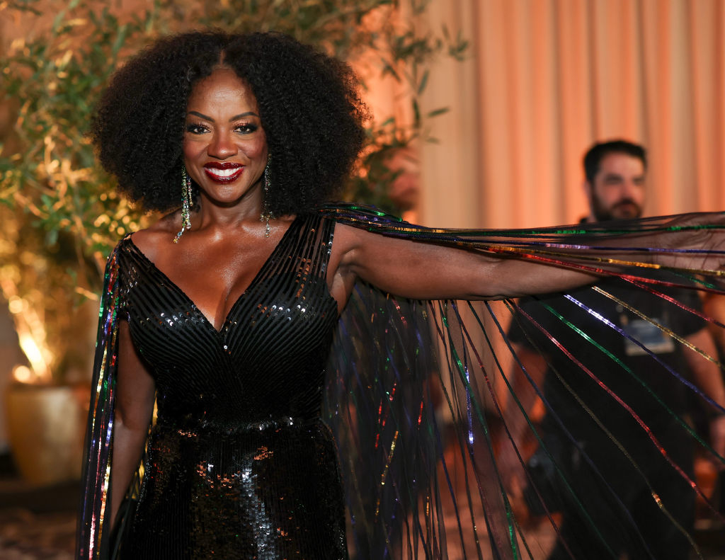 A woman in a sparkling black gown poses, smiling, with sheer, shimmering cape-like sleeves at a glamorous event