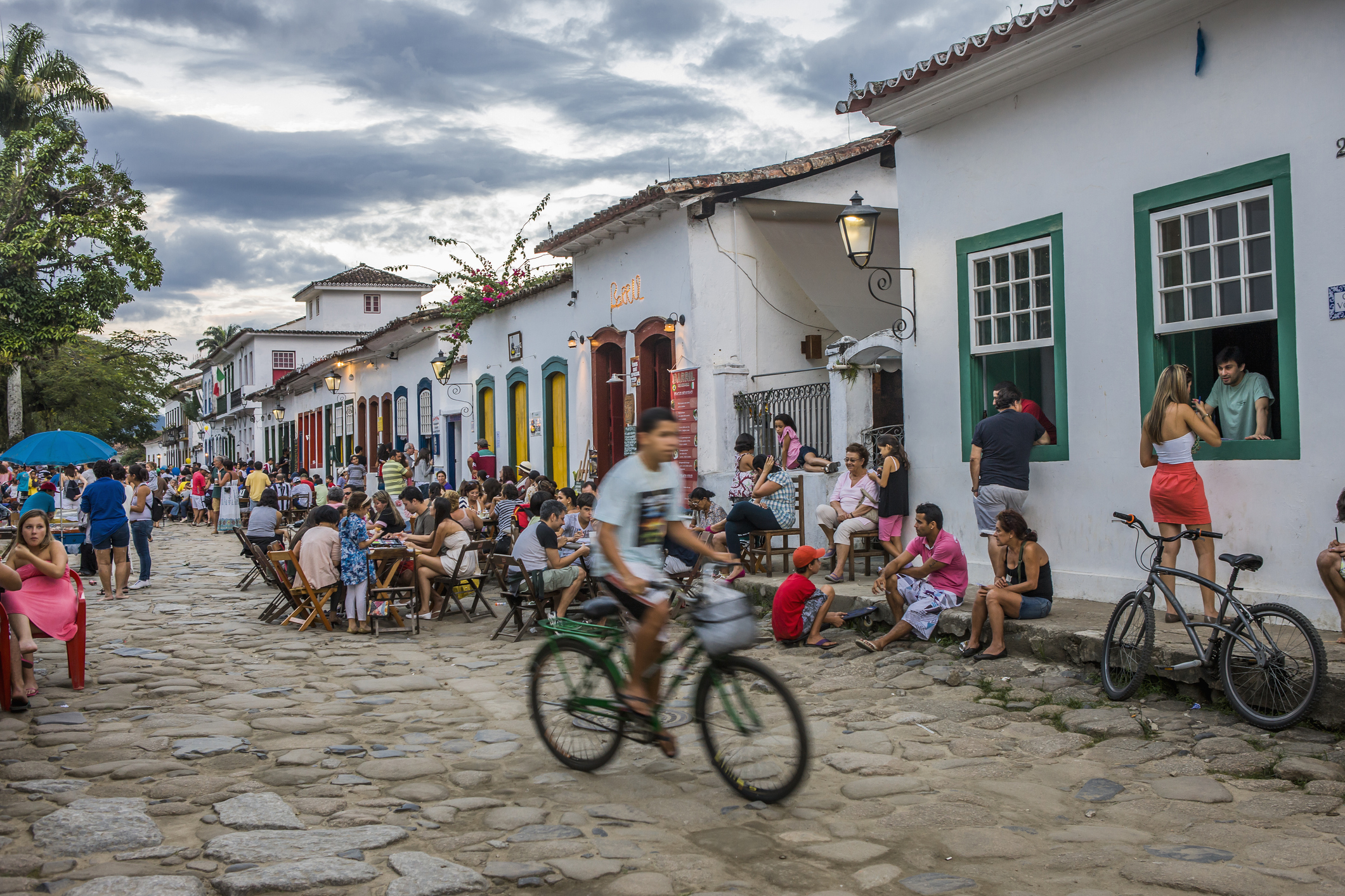 People sit along a cobblestone street lined with colorful buildings; a cyclist rides past