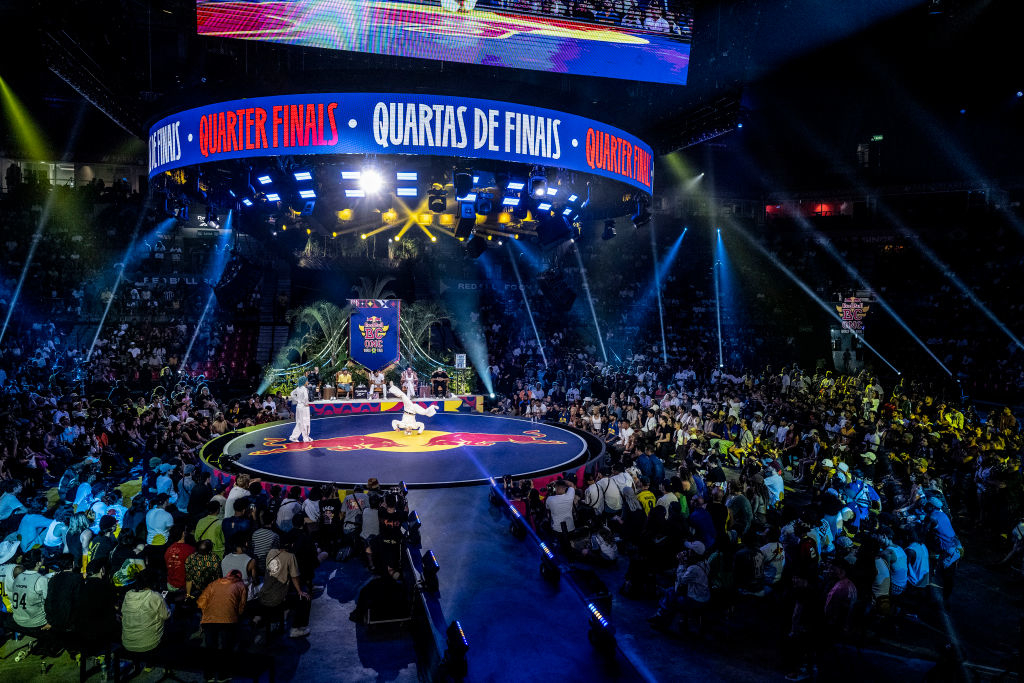 Breakdancing competition stage with an audience, large screen displaying &quot;Quarter Finals.&quot; Dancers performing on the circular stage