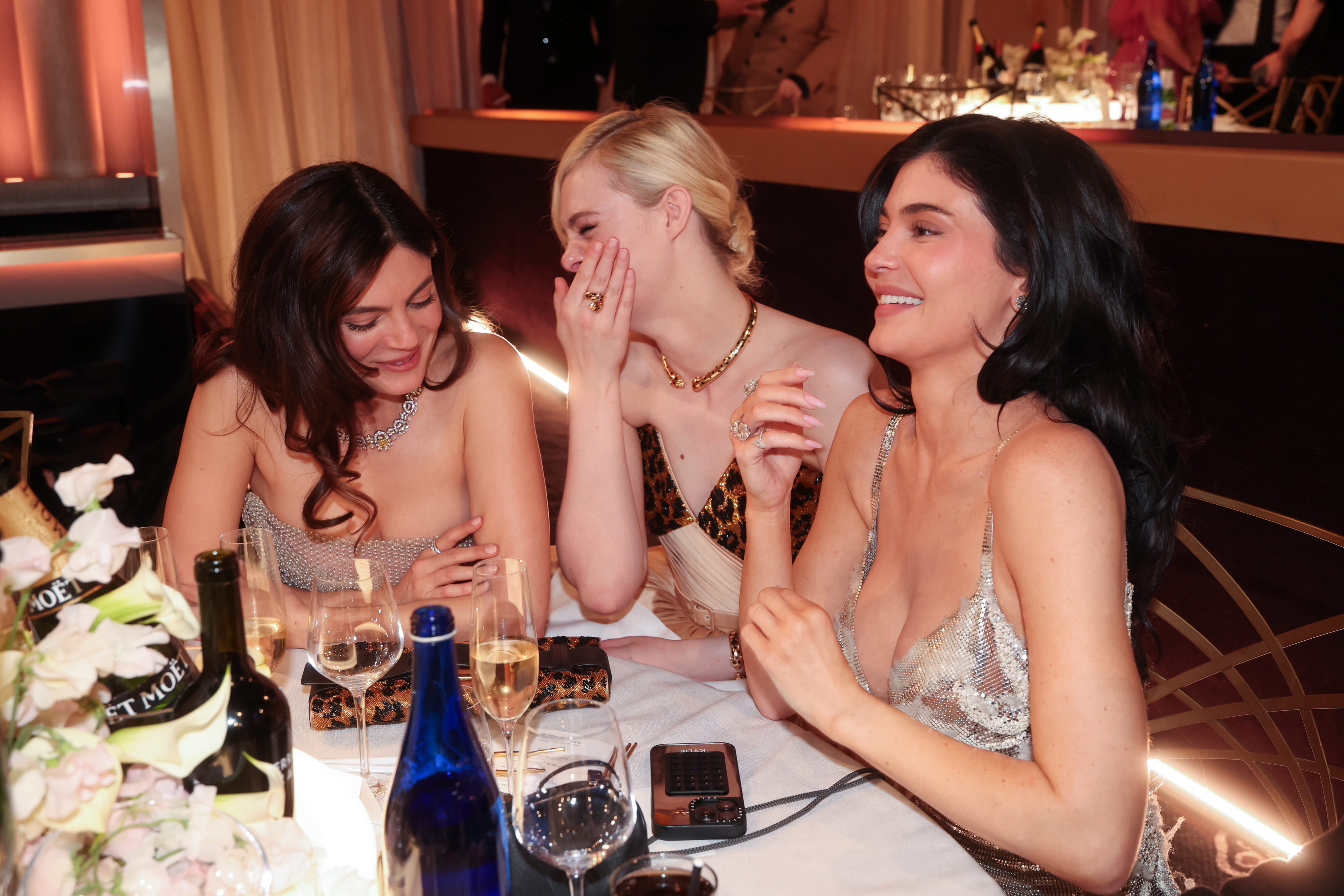 Three women in elegant evening gowns chat and laugh at a formal event table, surrounded by wine bottles and glasses