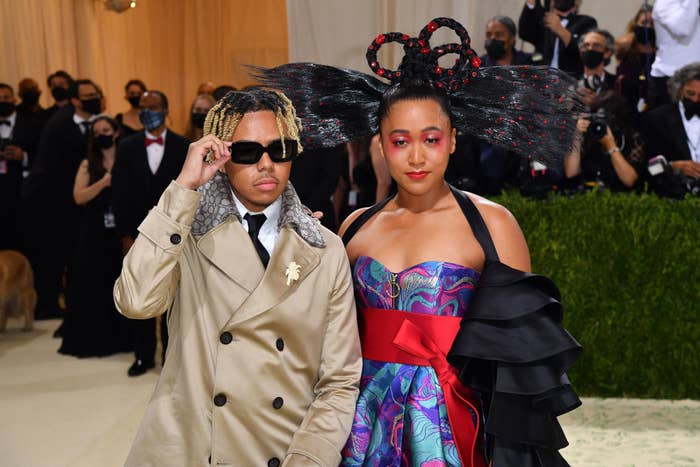 Cordae and Naomi Osaka on a formal event carpet. One wears a trench coat and sunglasses; the other wears a colorful strapless gown with elaborate hair styling